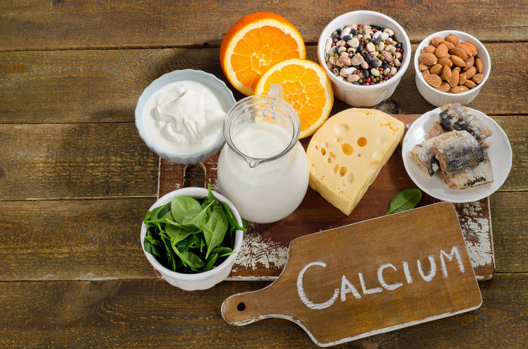 Calcium-rich foods on a wooden table including oranges, cheese, milk, leafy greens, nuts, and more. Next to a sign that says