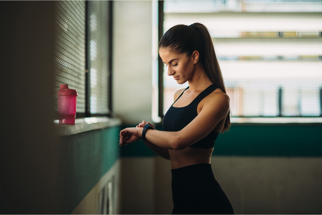Woman in workout gear checking fitness watch with a pink shaker bottle nearby in a gym setting.