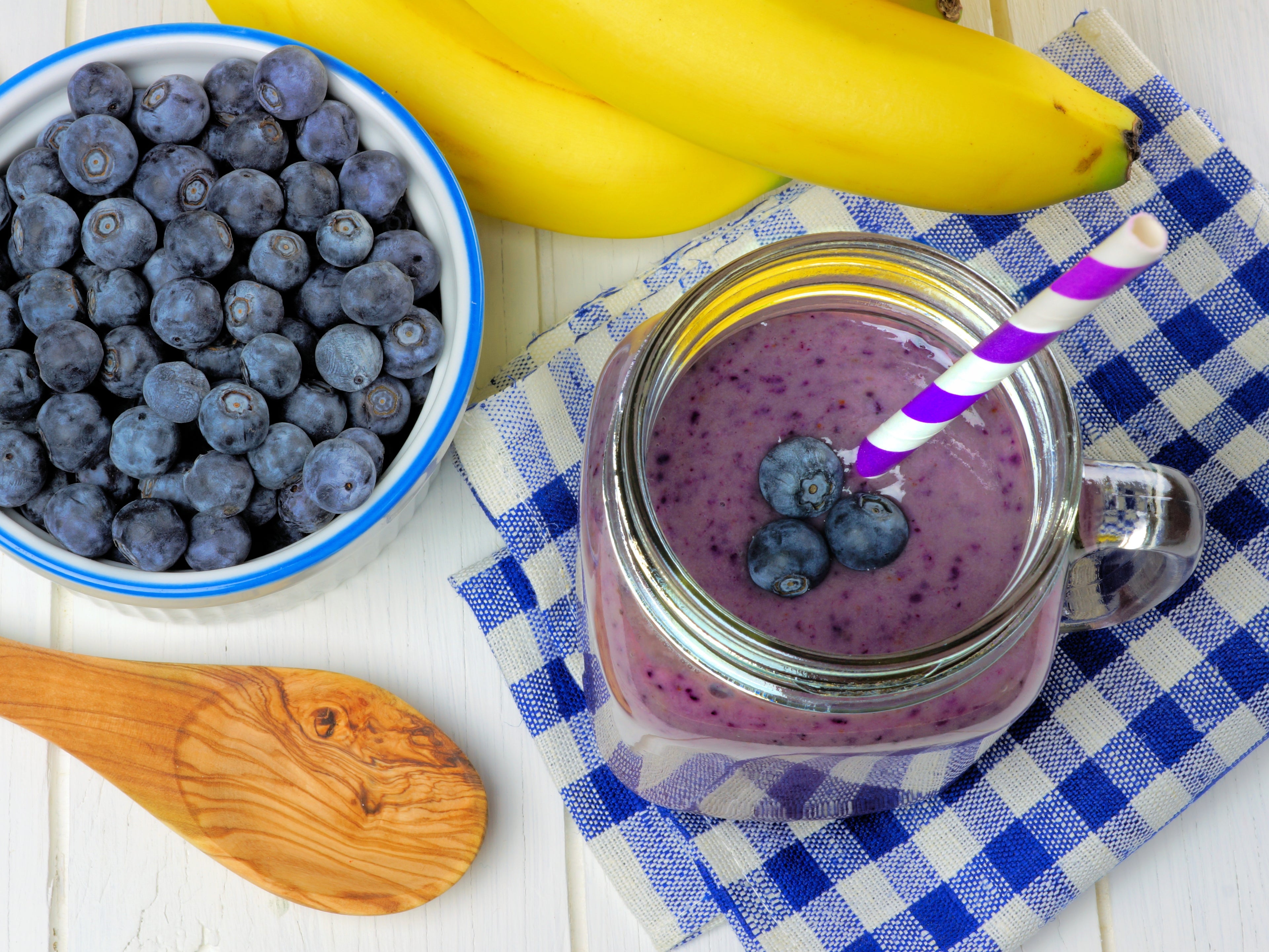 Blueberry smoothie above view with bananas checkered cloth and spoon on white wood