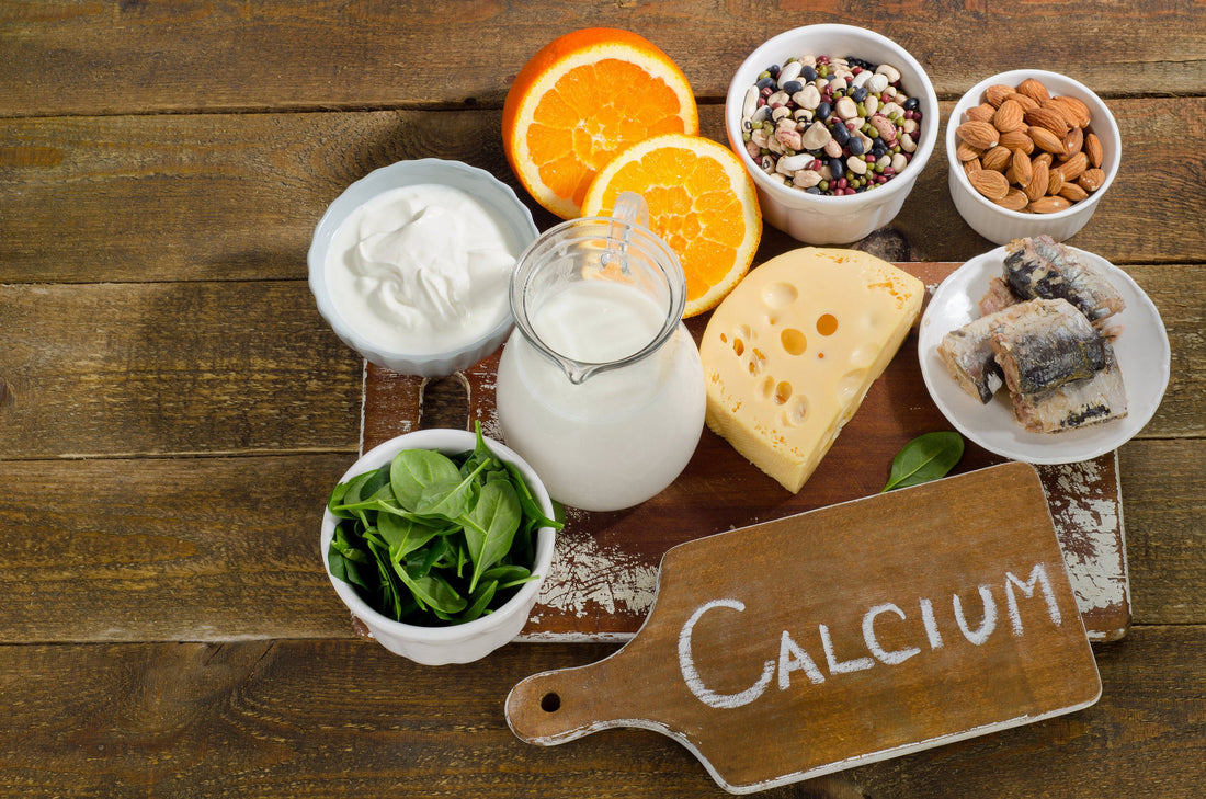 Calcium-rich foods on a wooden table including oranges, cheese, milk, leafy greens, nuts, and more. Next to a sign that says "calcium".