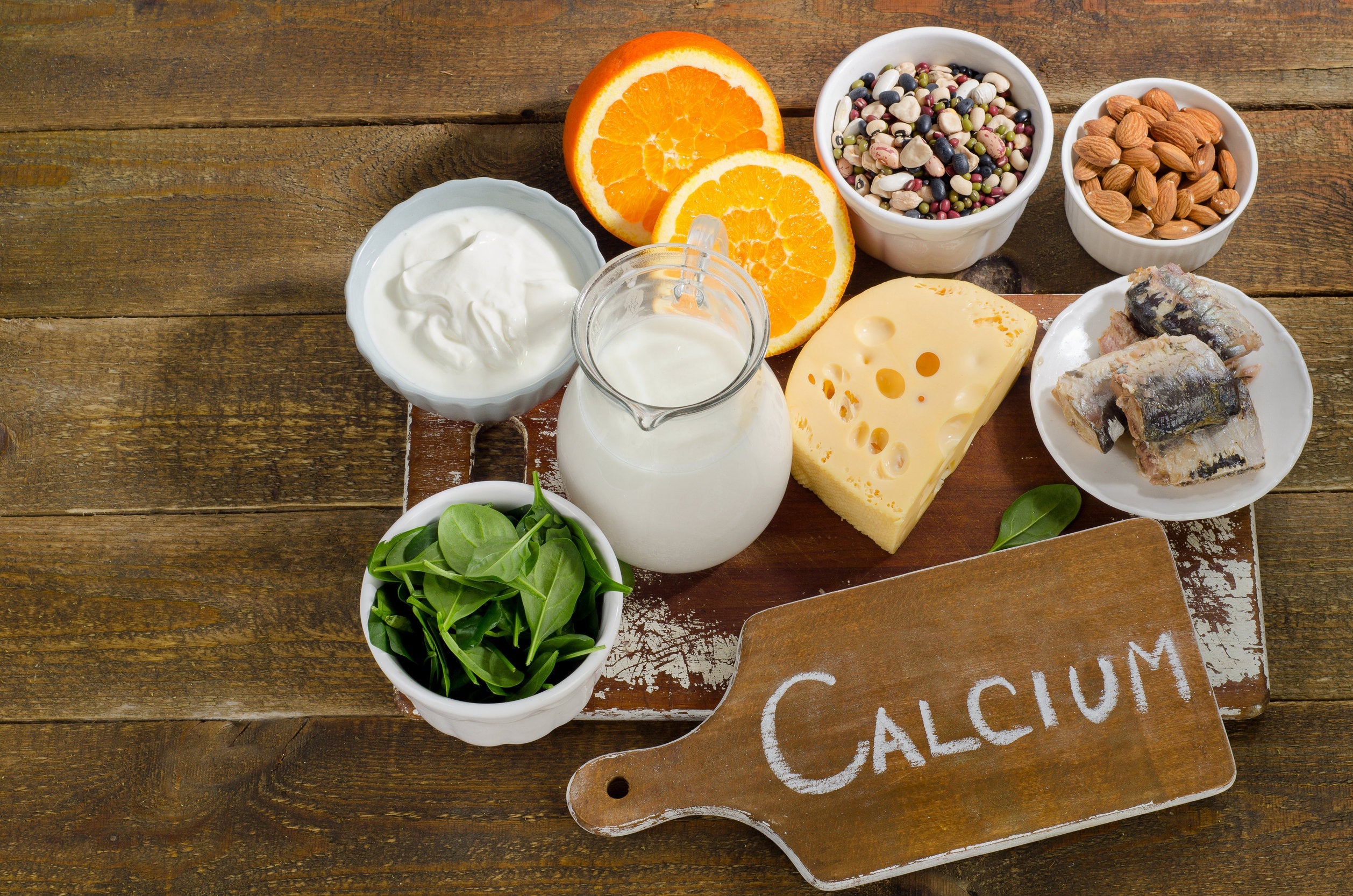 Calcium-rich foods on a wooden table including oranges, cheese, milk, leafy greens, nuts, and more. Next to a sign that says "calcium".