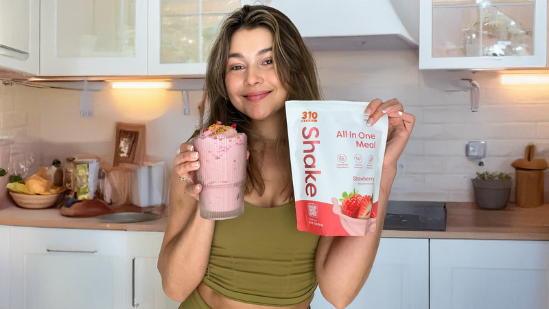 Woman holding a strawberry keto meal replacement shake and 310 Shake pouch in a bright kitchen.