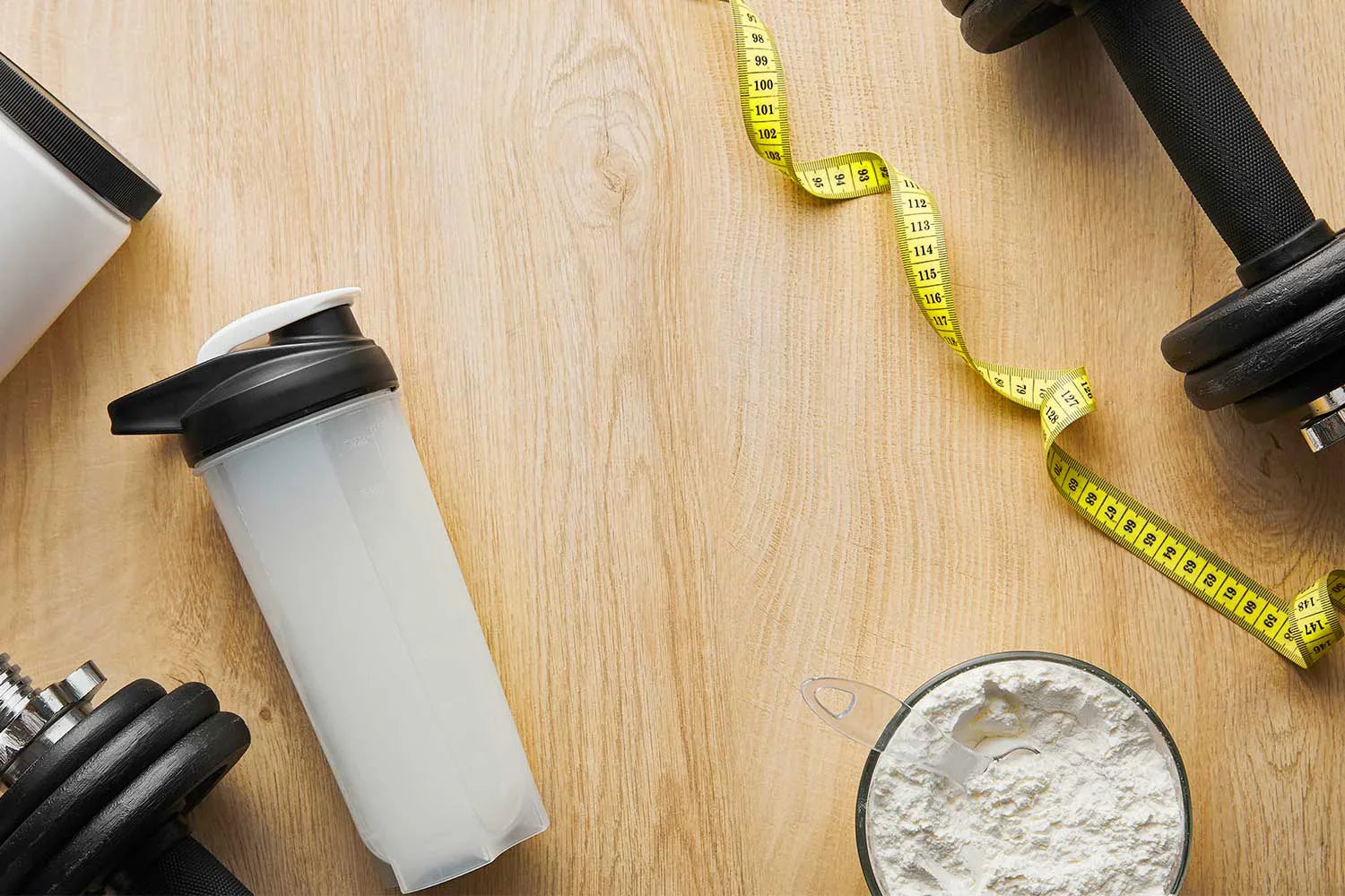 Dumbbells near a sports bottle and measuring tape on wooden surface