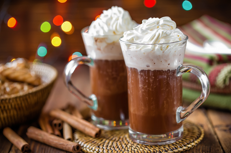 hot chocolate in glass mug with whipped cream, lights, and dark wood background