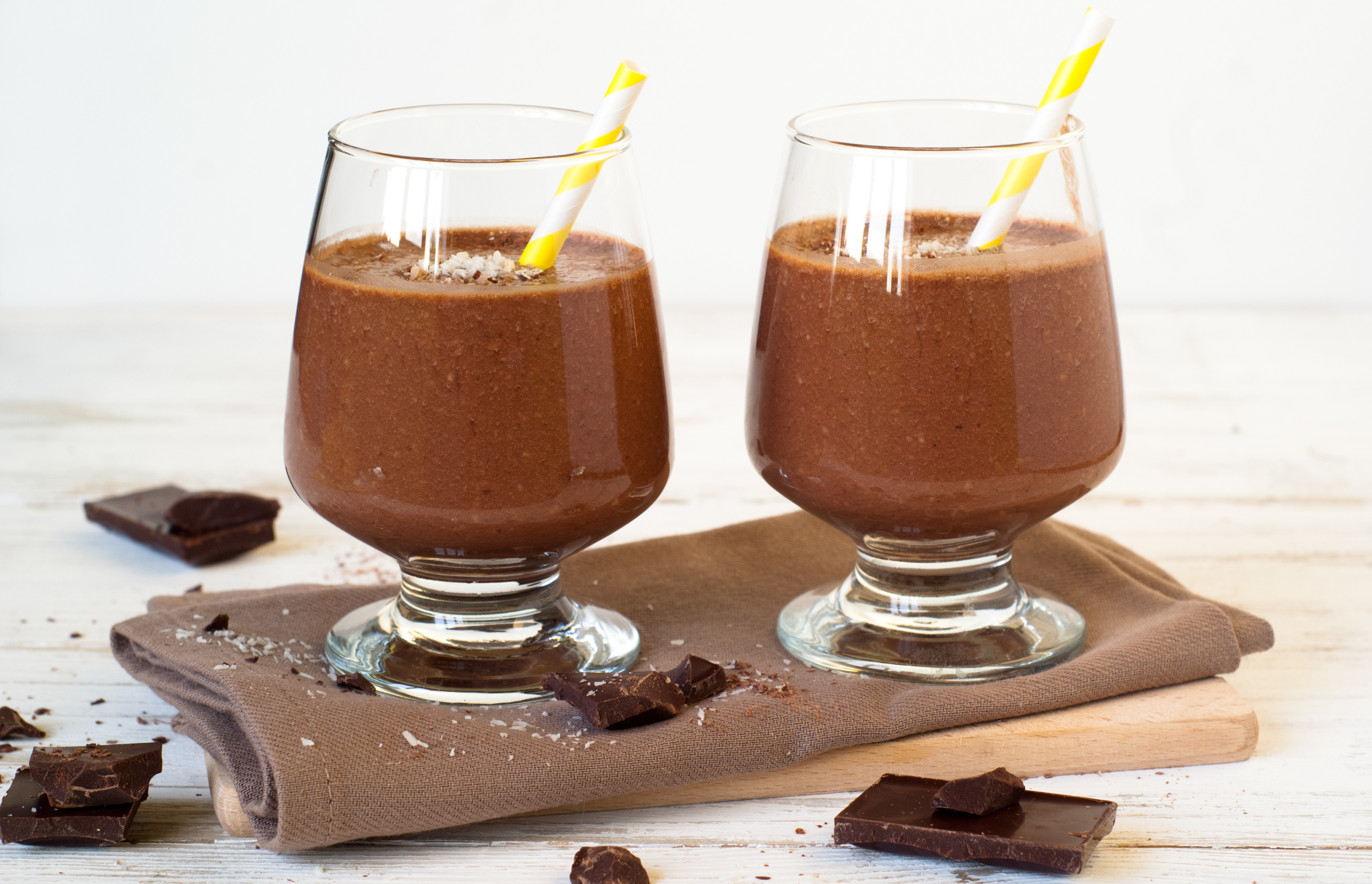 two glasses of chocolate coconut shake and chunks of chocolate on a wooden surface