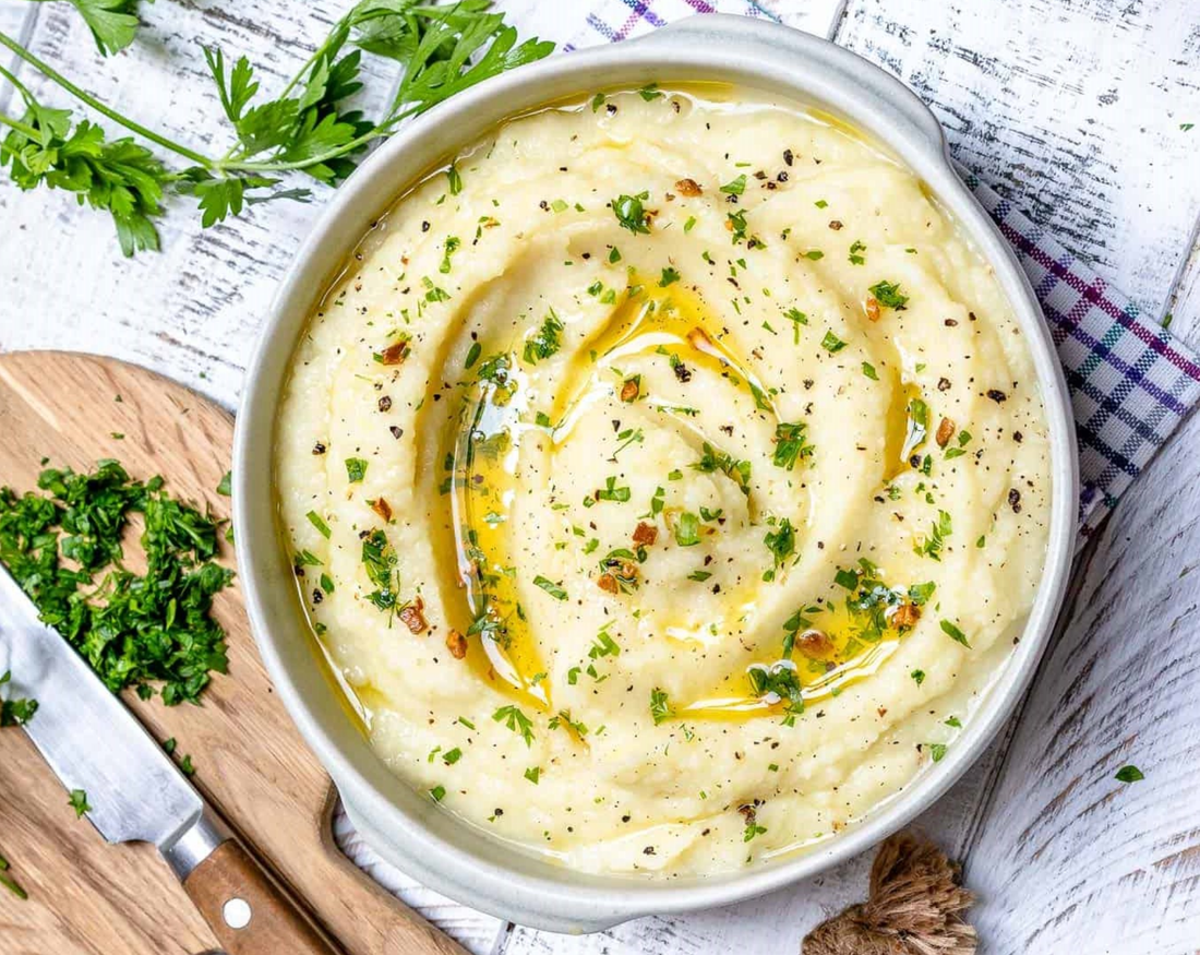 mashed cauliflower in a bowl with pasley and oil. Served on white wood with cutting board and knife