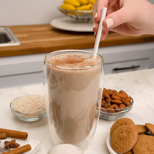 A glass of shake with a hand holding the straw, cookies, almonds, rice and cinnamon sticks.