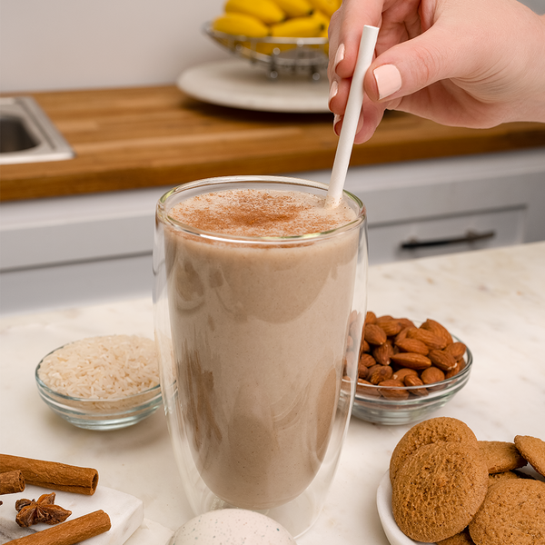 A glass of shake with a hand holding the straw, cookies, almonds, rice and cinnamon sticks.