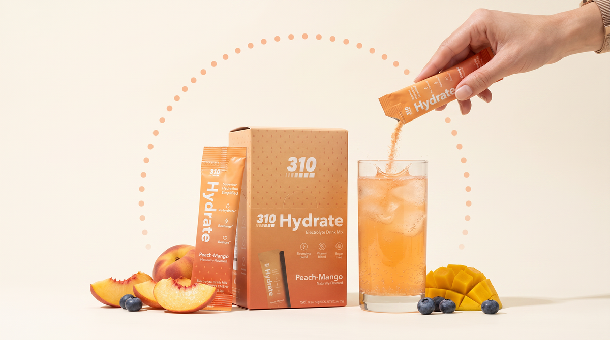 Pouring a 310 Hydrate Peach-Mango stick into a glass of iced electrolyte drink beside the 15-count box and fresh fruit