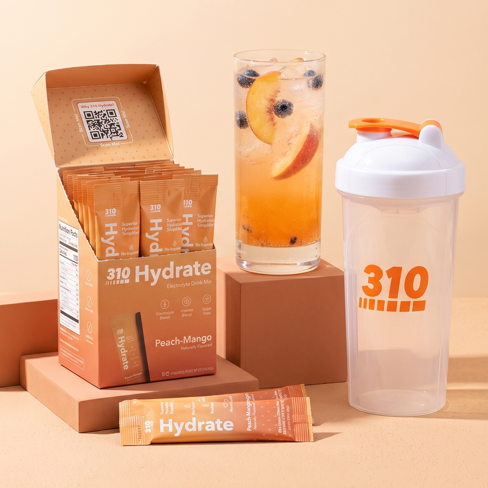 Open 310 Hydrate Peach Mango box with sticks, the 310 shaker cup and an iced peach drink on warm terracotta blocks