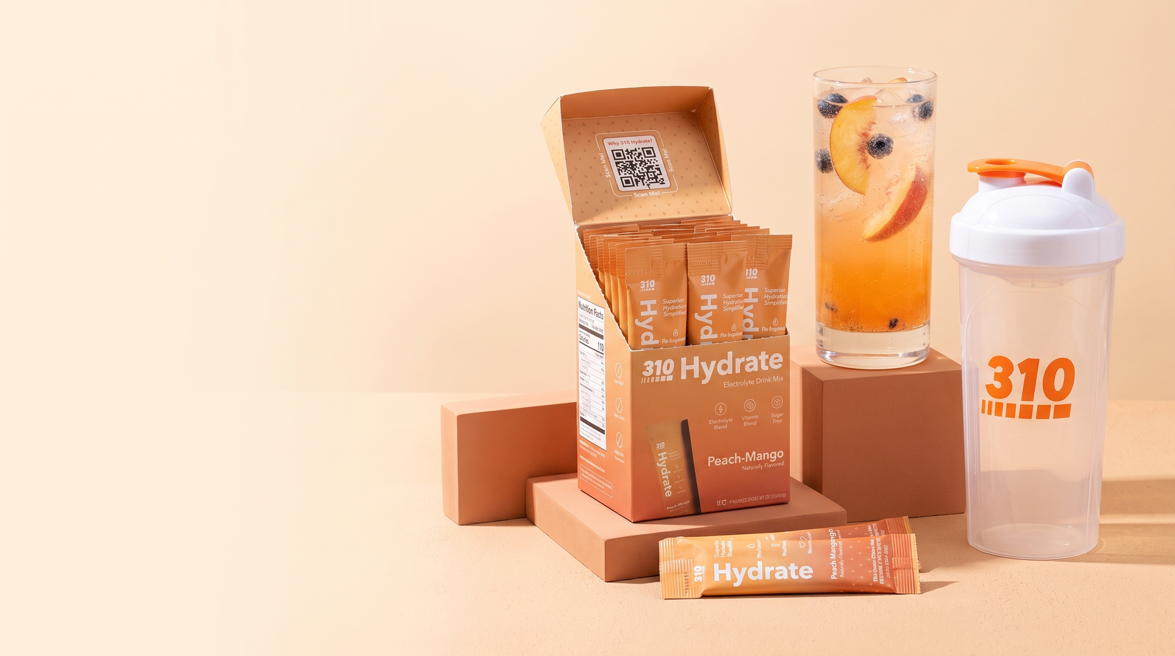 Open 310 Hydrate Peach Mango box with sticks, the 310 shaker cup and an iced peach drink on warm terracotta blocks