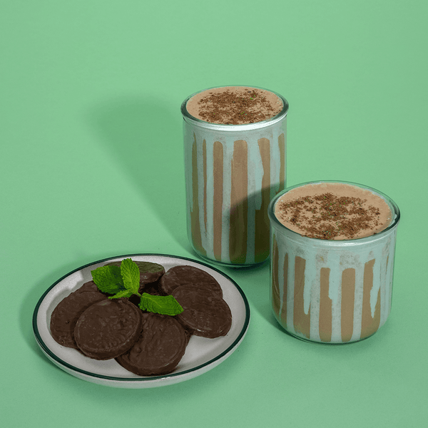 Two glasses of 310 Chocolate Mint protein shake and a plate of treats in a mint-themed background.